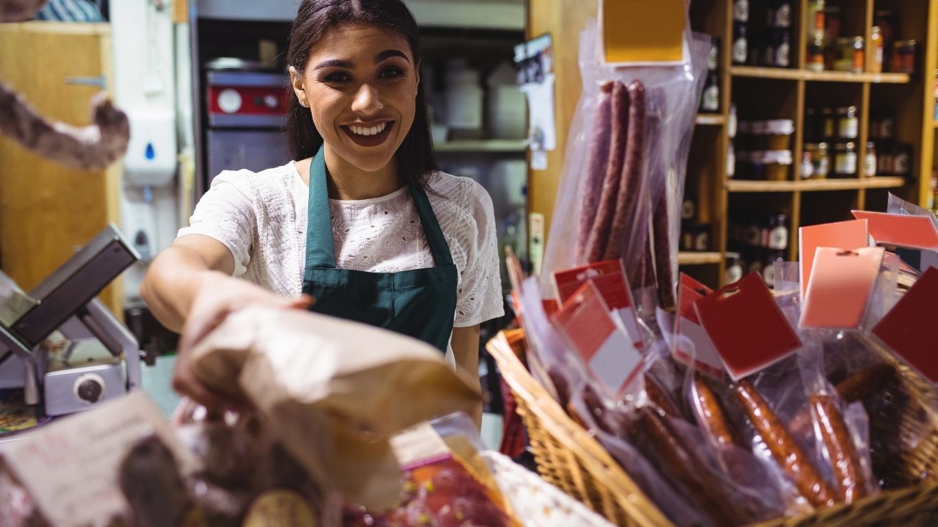 Mujer mexicana sonriente atendiendo su propio negocio de abarrotes con estantes llenos de mercancía.